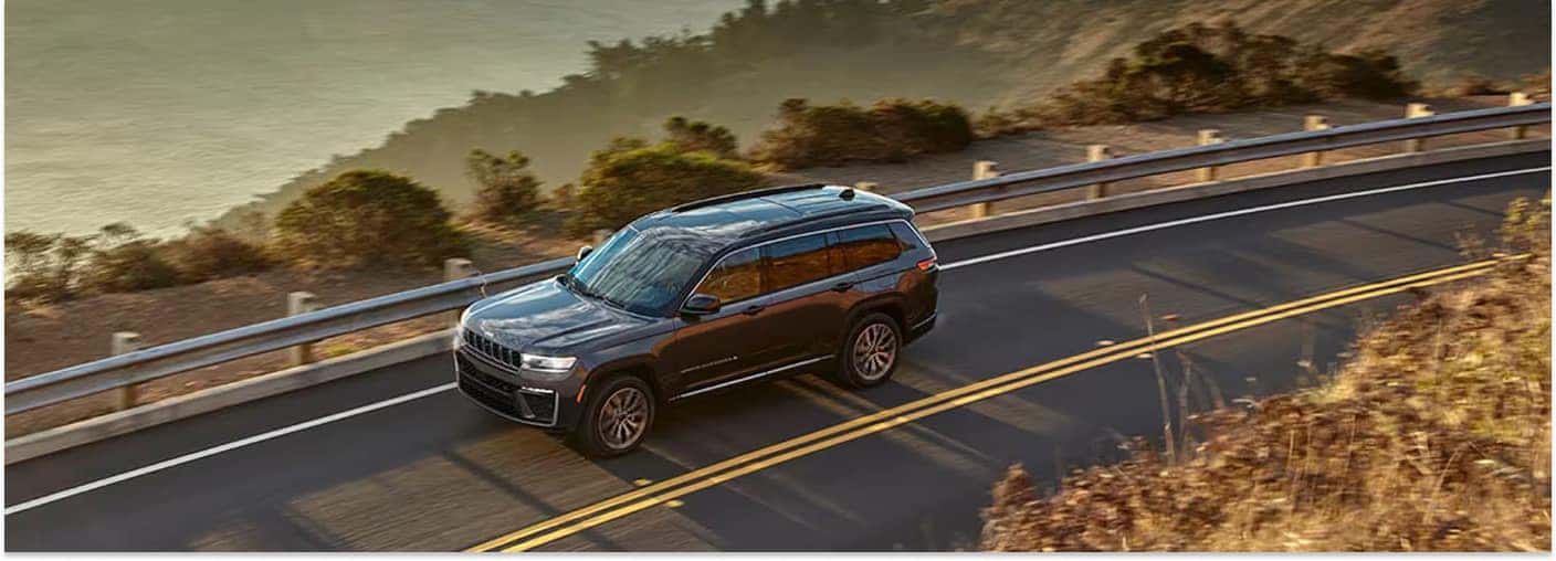 > High-angle view of a dark gray 2026 Jeep Grand Cherokee L driving along a winding coastal highway bordered by a guardrail under warm, late-afternoon sunlight.