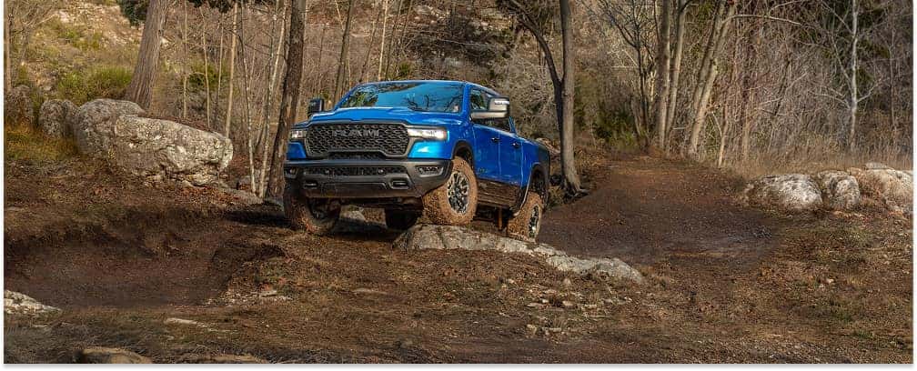 Blue 2026 RAM 1500 pickup truck driving on a dirt trail in a wooded area, shown from a front three-quarter view.