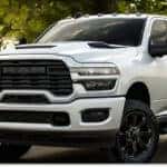 > Front three-quarter view of a white 2026 RAM 2500 heavy-duty pickup truck with a bold black grille and matching black wheels, parked on an asphalt surface against a lush, tree-lined background.