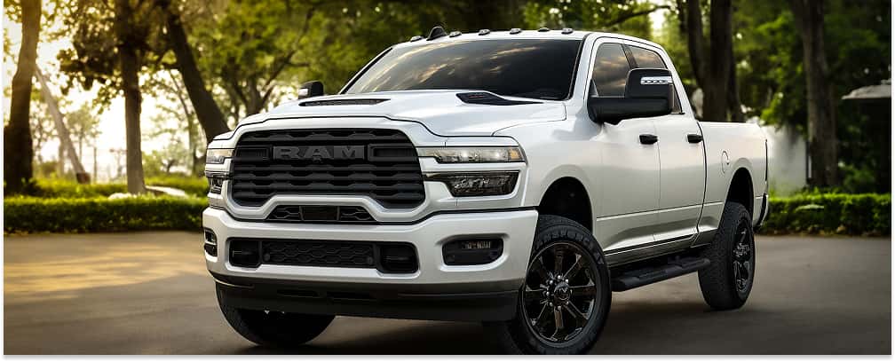 > Front three-quarter view of a white 2026 RAM 2500 heavy-duty pickup truck with a bold black grille and matching black wheels, parked on an asphalt surface against a lush, tree-lined background.