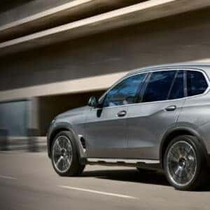 Side and rear view of a silver 2026 BMW X5 xDrive driving through a modern city environment