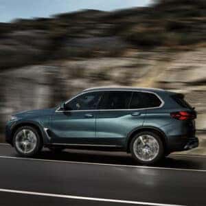Side view of a 2026 BMW X5 in motion on a scenic road with a rocky background
