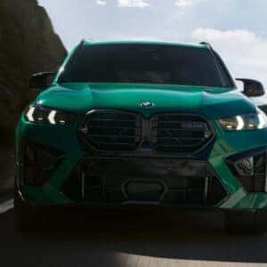 Front view of a green 2026 BMW X5 M driving through a mountain pass