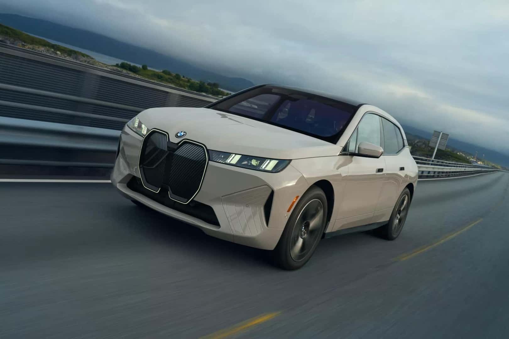 a cream colored bmw ix driving on a highway in cloudy weather with a city and body of water in the background