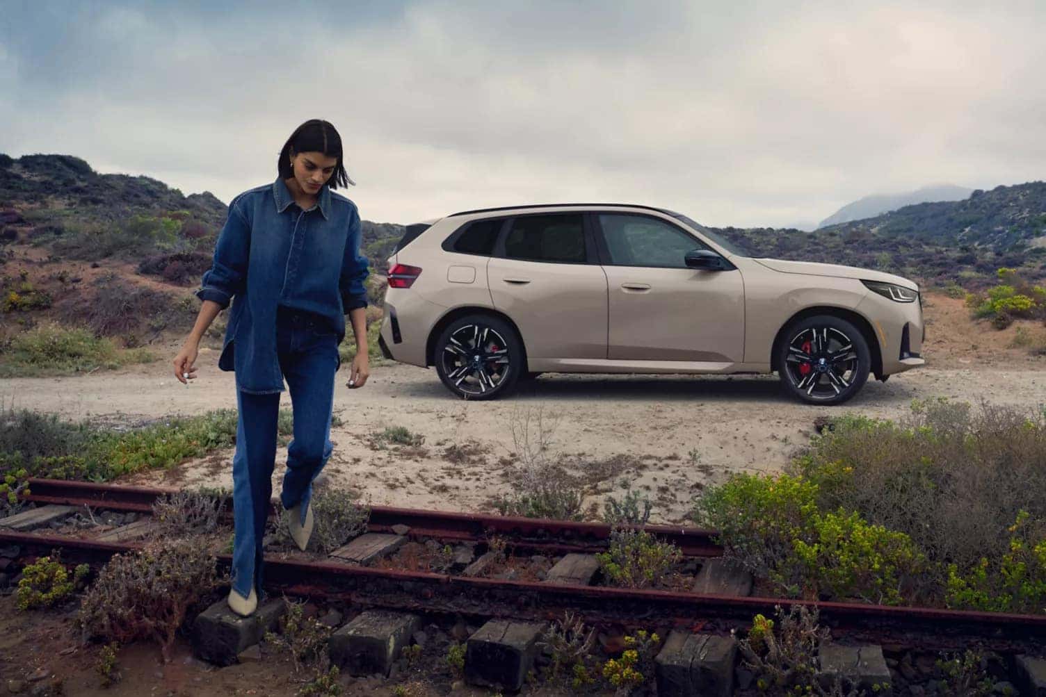 a woman walking on train tracks and away from a 2026 bmw x3 parked on a gravel path
