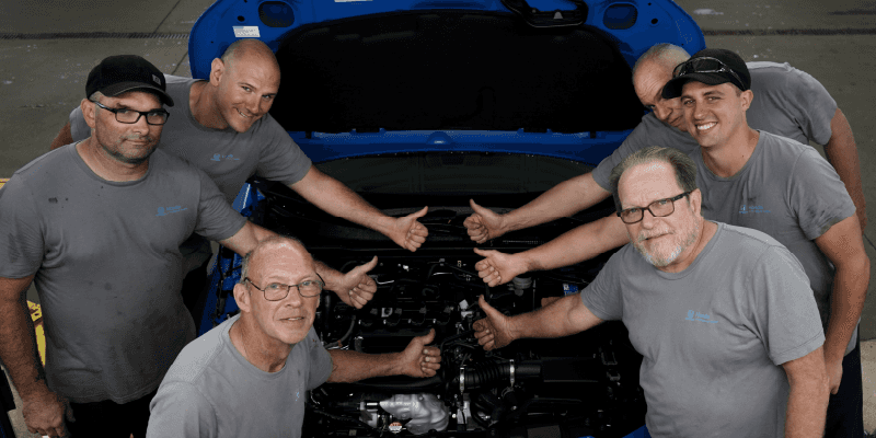 Techs in front of a car engine giving the thumbs up.