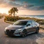 A gray Nissan Sentra parked on a coastal road at sunset with a beach and palm trees in the background.