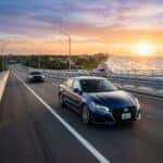 A dark blue Nissan Altima driving on a coastal highway at sunset with the ocean in the background.