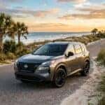 A gray Nissan Rogue parked on a coastal road during sunset with palm trees and beach in the background.
