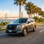 Grey Nissan SUV driving along a coastal road lined with palm trees near a large suspension bridge.