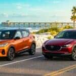 An orange Nissan Kicks and a red Mazda CX-30 parked on a coastal road near a beach and pier.