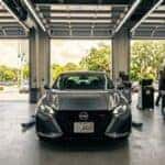 Front view of a gray Nissan Altima inside a bright automotive service bay with mechanics working in the background.