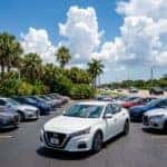 A white sedan parked on a car dealership lot under a bright blue sky with fluffy clouds and palm trees.