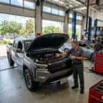 A Nissan technician points to the engine of a grey Frontier pickup truck in a service garage.