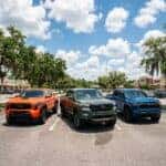 Three mid-size pickup trucks in orange, green, and blue parked side-by-side in a sunny parking lot.