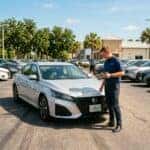 A dealership employee in a blue shirt holds a tablet while inspecting a silver Nissan sedan parked on a sunny lot.