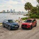 A blue Nissan Rogue and red Honda CR-V parked in a lot near a riverfront with a couple walking in the background. (Representative image for illustration purposes only. Actual vehicle may vary based on trim and configuration.)