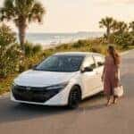 Woman in a pink dress walking past a white sedan on a coastal road near the ocean. (Representative image for illustration purposes only. Actual vehicle may vary based on trim and configuration.)