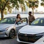 A woman and man talk between a silver Hyundai Elantra and a white Nissan Sentra parked near a waterfront park. (Representative image for illustration purposes only. Actual vehicle may vary based on trim and configuration.)