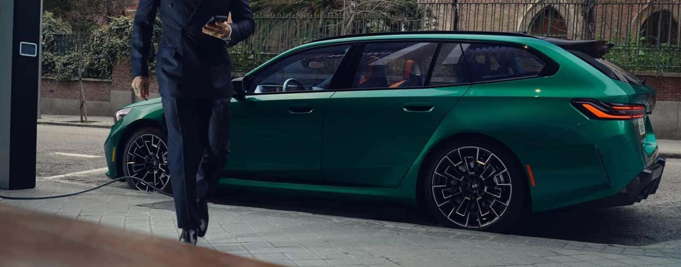 A green 2025 BMW M5 Touring is parked on the side of a street while charging.