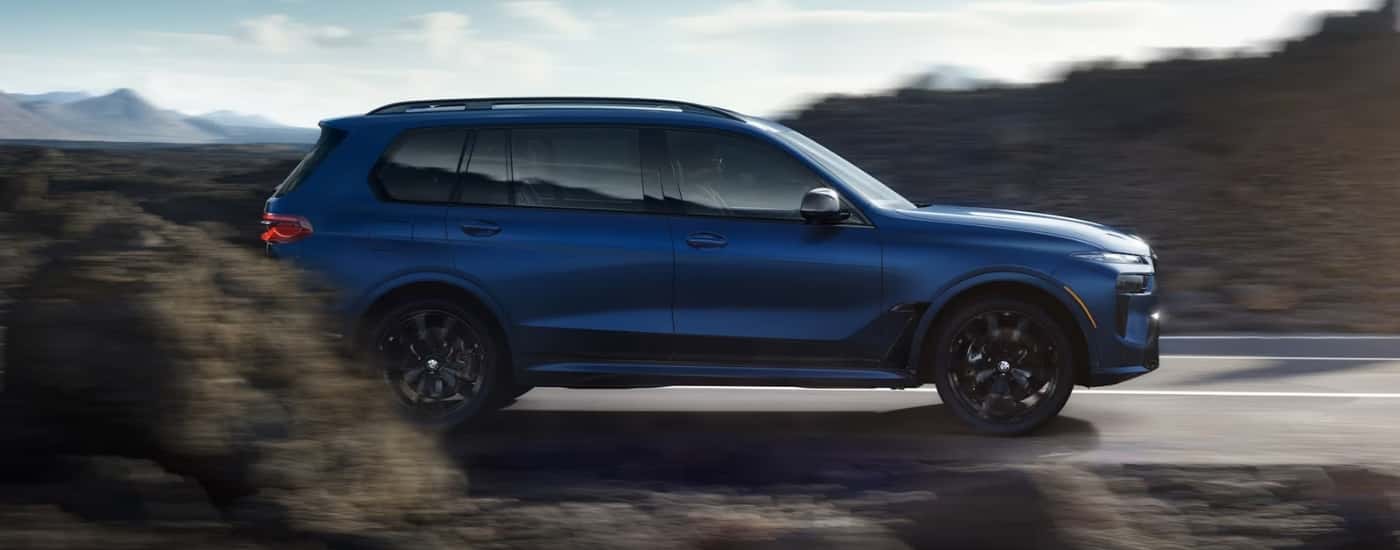 A blue 2025 BMW X7 M60i speeding on a small road.
