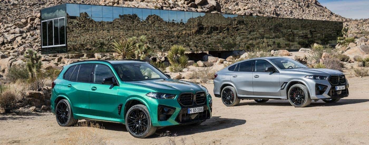 A teal 2026 BMW X5M and a silver 2026 BMW X6M parked.