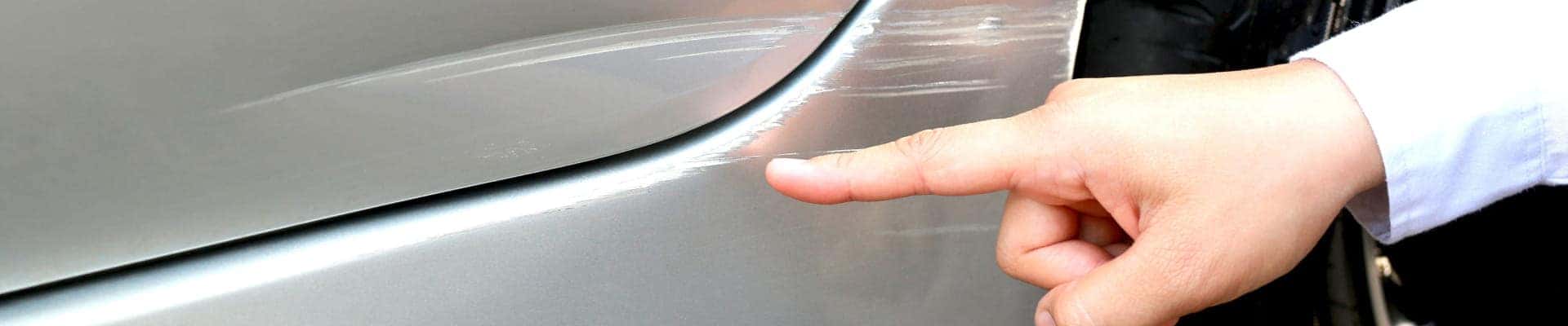 A hand points to a scratch on a silver car's surface, highlighting the damage. The focus is on the visible scrape, conveying inspection.