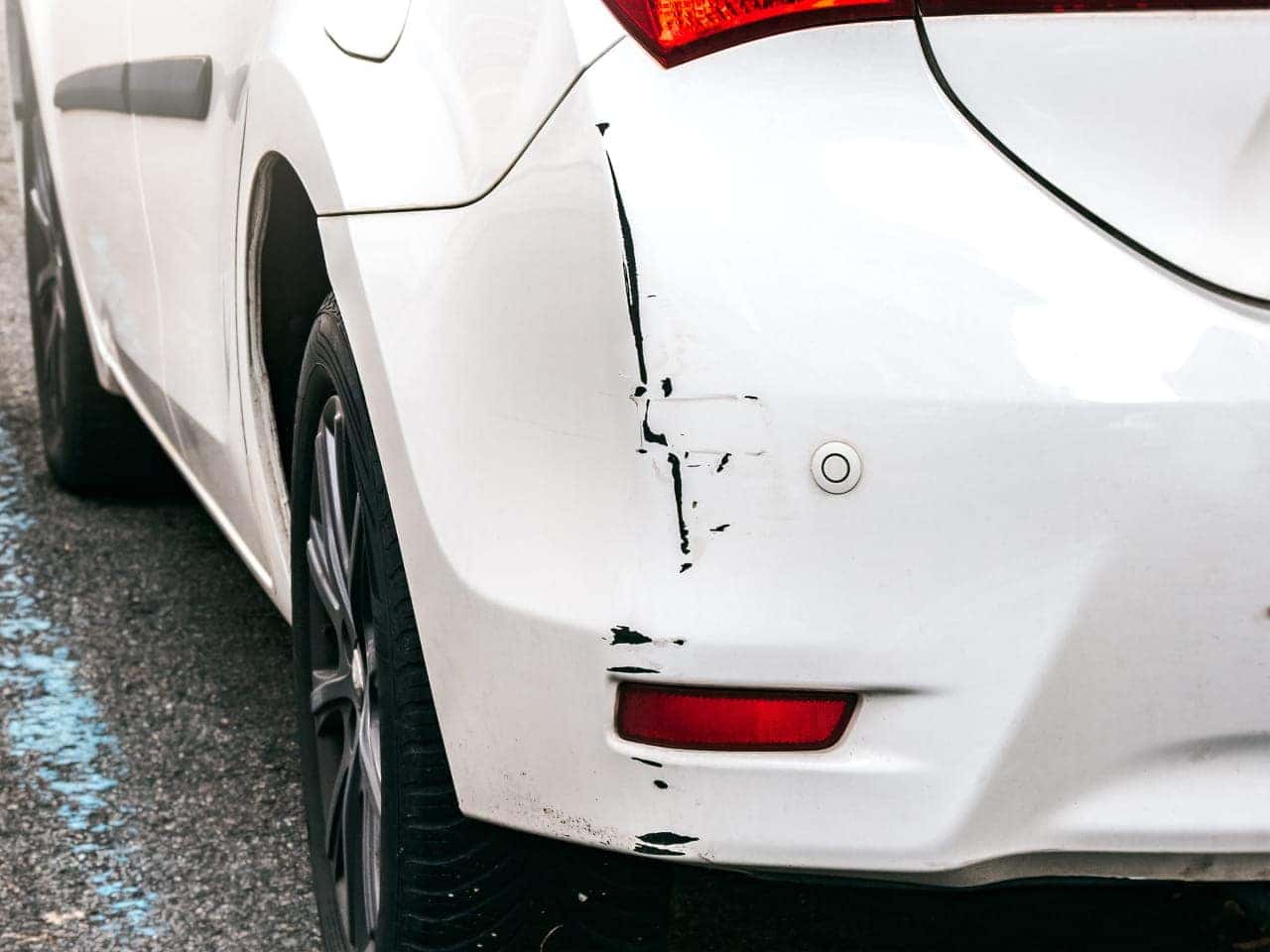 Close-up of a white car's rear bumper with noticeable scratches and black marks, indicating minor damage.
