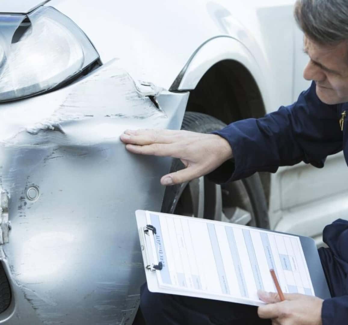 A man with a clipboard inspects a severe dent in a car's front bumper.