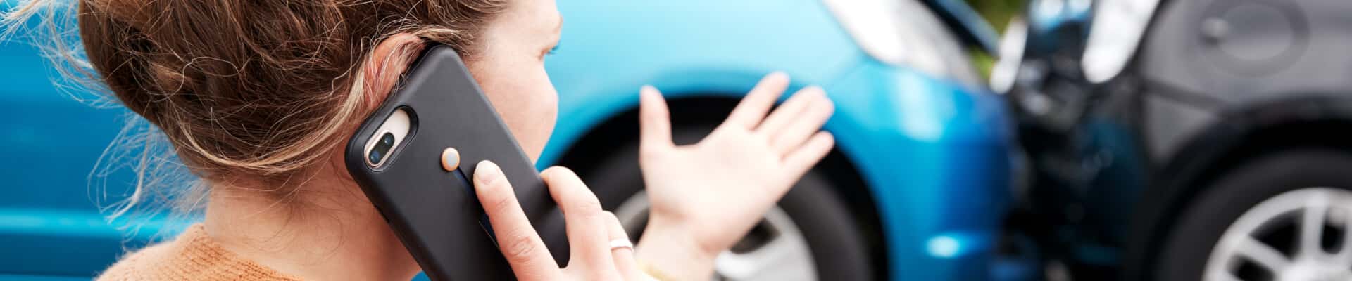 A woman holds a cell phone to her ear while gesturing towards a car accident in the background.