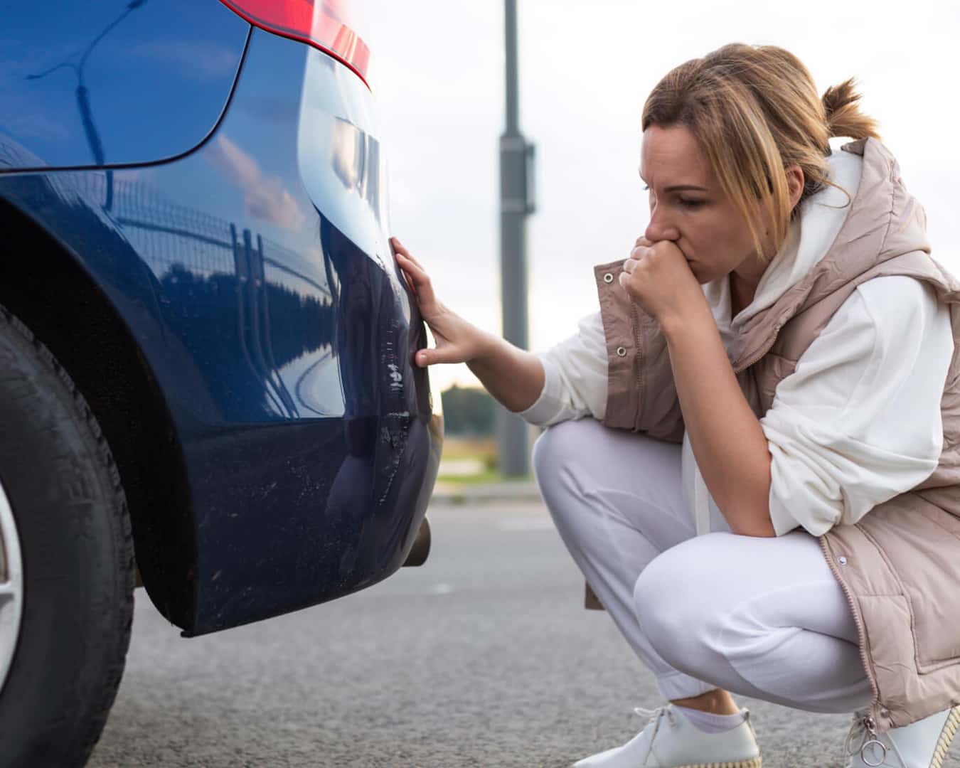 A close-up of a woman kneeling to assess the damage to the bumper of a blue car.