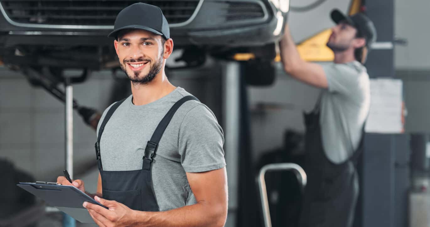 A mechanic holding a clipboard smiles as another mechanic works on a vehicle on a lift.